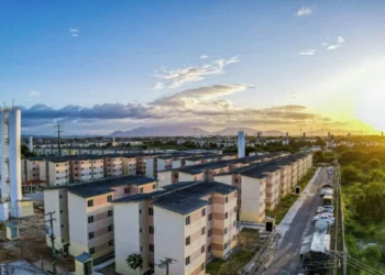 Vista aérea de conjunto habitacional do programa Minha Casa Minha Vida, com várias unidades residenciais e a luz do pôr do sol ao fundo.
