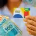 Pessoa segurando o cartão do Bolsa Família com notas de real ao fundo