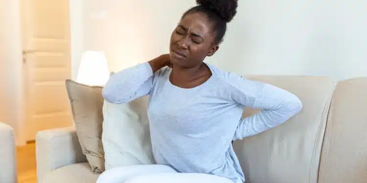 Jovem mulher negra com dor nas costas devido à fibromialgia, em casa, sentada no sofá.