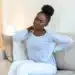 Jovem mulher negra com dor nas costas devido à fibromialgia, em casa, sentada no sofá.