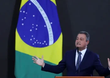 Ministro Rui Costa discursando sobre a desvinculação do salário mínimo da aposentadoria, com a bandeira do Brasil ao fundo.