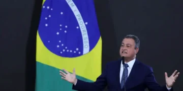 Ministro Rui Costa discursando sobre a desvinculação do salário mínimo da aposentadoria, com a bandeira do Brasil ao fundo.