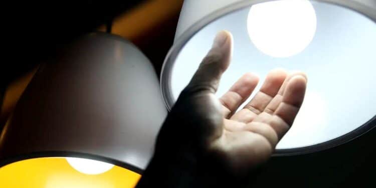 Mão de uma pessoa estendida abaixo de uma lâmpada acesa. A imagem simboliza o alto custo da energia elétrica e o impacto da bandeira vermelha 2 nas contas de luz.