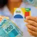 Cartão do Bolsa Família sendo segurado por uma mão, com notas de dinheiro do Brasil em primeiro plano.