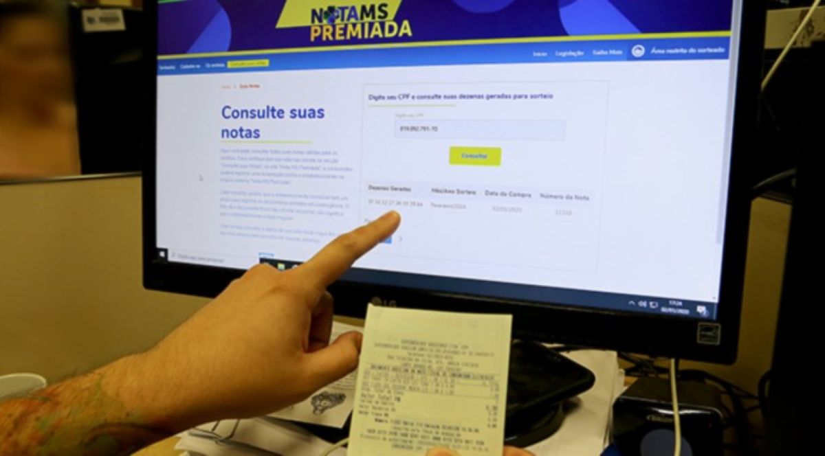 Consulta de notas premiadas no site da Nota Premiada MS com um recibo em mãos.