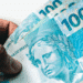 Notas de R$ 100 do Brasil sendo seguradas, relacionadas ao crédito de R$ 150 liberado pela Caixa Econômica.