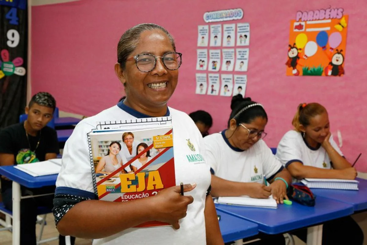Mulher sorrindo com um livro nas mãos, dentro de uma sala de aula de Educação de Jovens e Adultos (EJA).