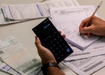 Pessoa usando calculadora no celular para organizar contas e boletos em cima da mesa.