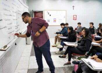 Professor escrevendo na lousa em uma sala de aula, ensinando a um grupo de alunos. A imagem representa a valorização da educação e o tema da Carteira Nacional do Professor.