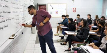 Professor escrevendo na lousa em uma sala de aula, ensinando a um grupo de alunos. A imagem representa a valorização da educação e o tema da Carteira Nacional do Professor.