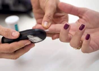 Um profissional de saúde realizando um teste de glicemia no dedo de uma pessoa, usando um glicosímetro. A imagem representa o monitoramento do diabetes e a atenção à saúde de pessoas com essa condição.