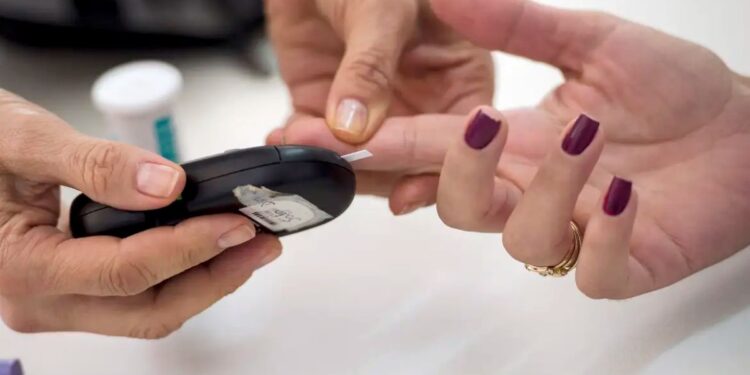 Um profissional de saúde realizando um teste de glicemia no dedo de uma pessoa, usando um glicosímetro. A imagem representa o monitoramento do diabetes e a atenção à saúde de pessoas com essa condição.