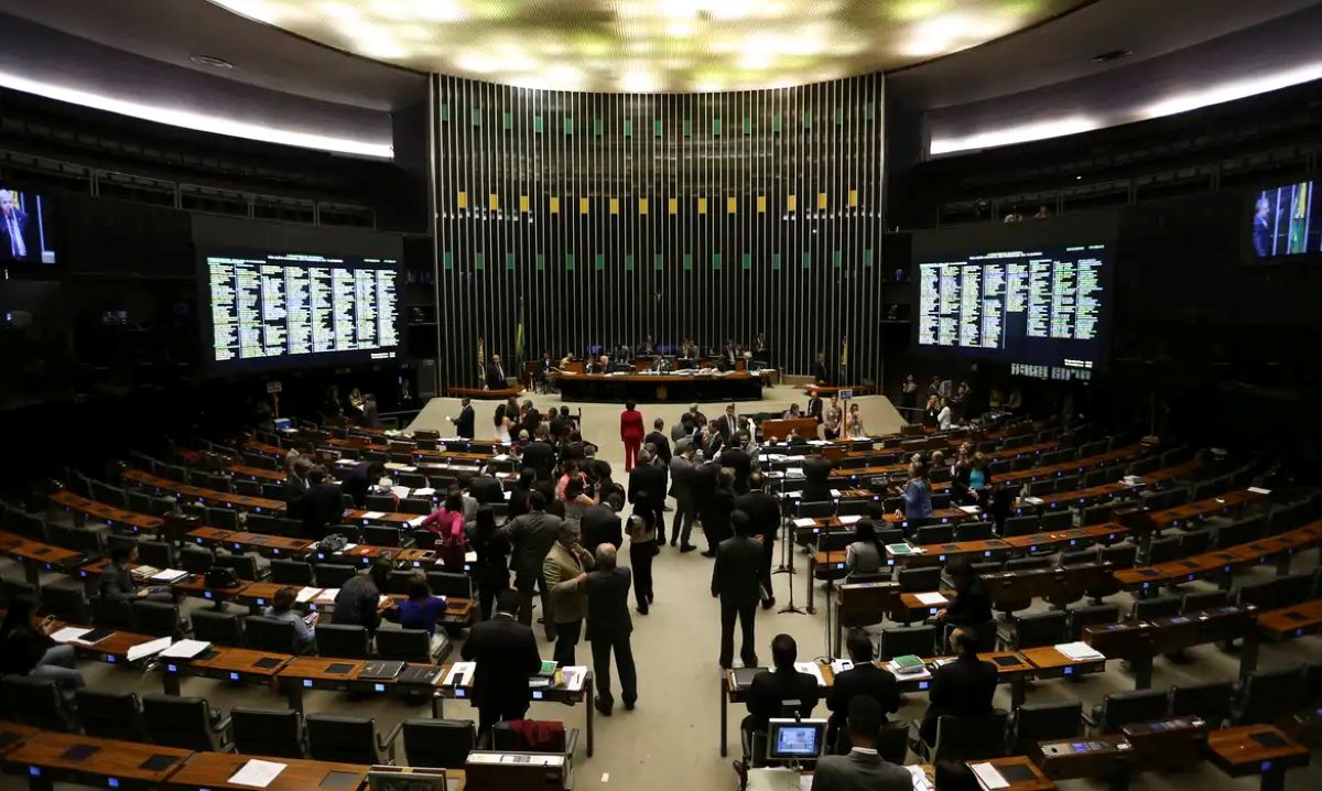 Plenário do Congresso Nacional, com parlamentares e público, representando o local onde as decisões sobre o salário mínimo e outros reajustes são tomadas, com displays digitais mostrando informações.