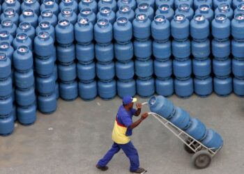 Homem uniformizado empurra um carrinho com vários botijões de gás azul e branco, em frente a uma pilha enorme desses botijões.