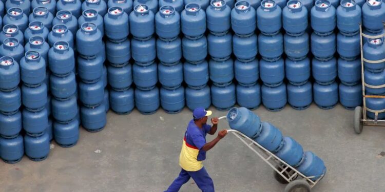 Homem uniformizado empurra um carrinho com vários botijões de gás azul e branco, em frente a uma pilha enorme desses botijões.