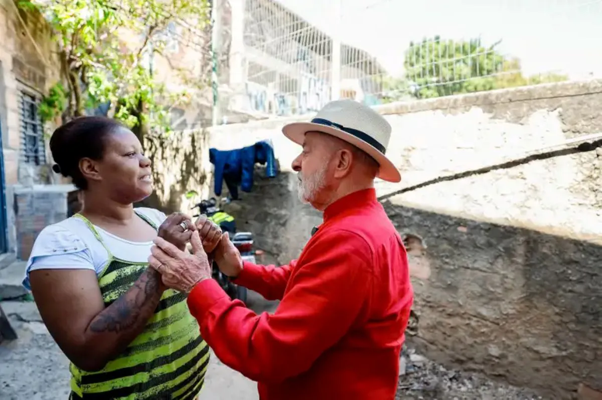 O presidente Lula, de camisa vermelha e chapéu de palha, segura as mãos de uma mulher com camisa listrada e conversa com ela em uma comunidade de baixa renda.