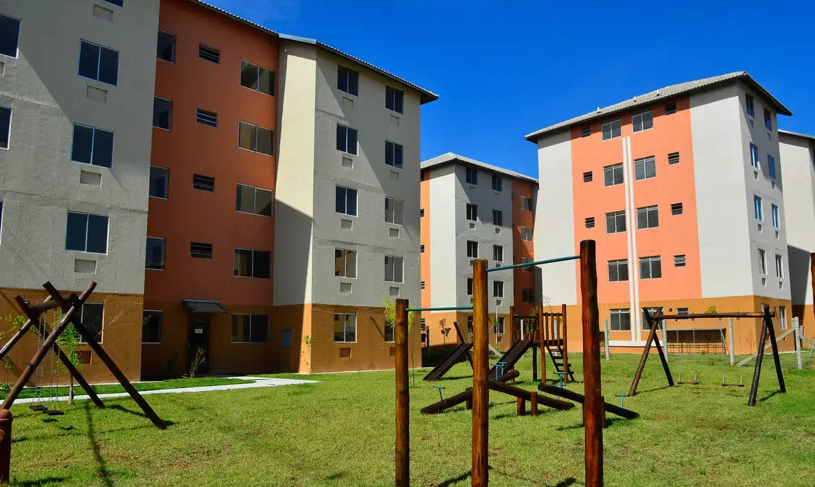 Edifícios coloridos de um condomínio residencial com um parque infantil de madeira e gramado verde sob um céu azul, representando moradias acessíveis.