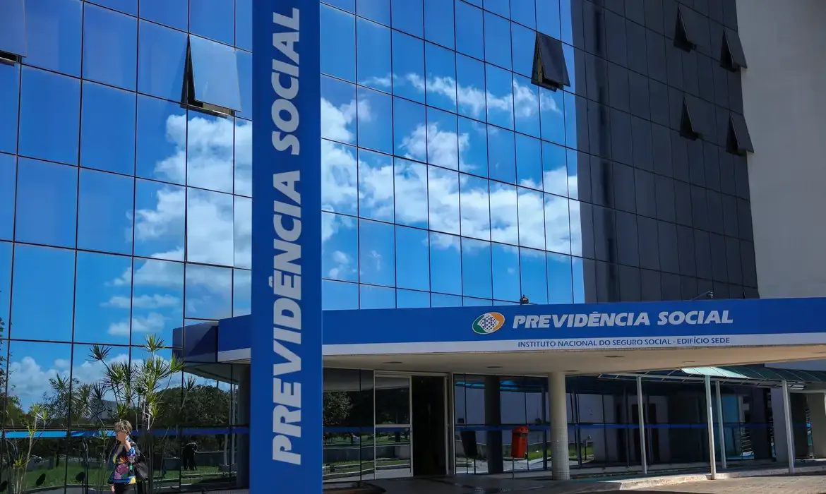 Agência do INSS com fachada de vidro refletindo o céu, localizada em um ambiente urbano.