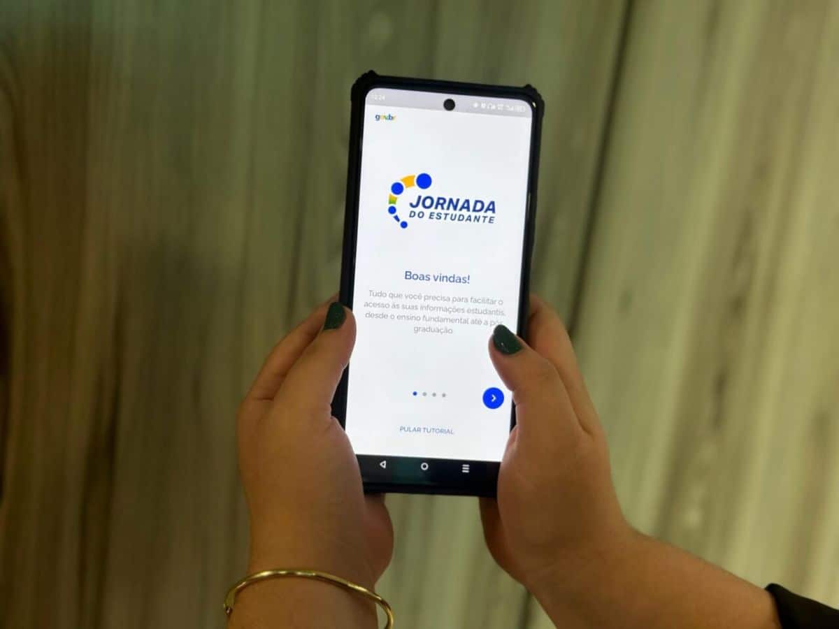 Mãos segurando um smartphone com o aplicativo "Jornada do Estudante" aberto, a ferramenta oficial para consultar pagamentos e informações sobre o programa Pé-de-Meia.
