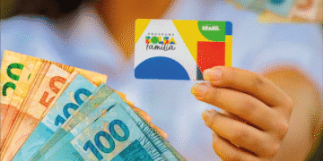 Pessoa segurando cartão do Programa Bolsa Família com notas de 100 reais ao fundo