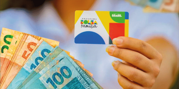 Pessoa segurando cartão do Programa Bolsa Família com notas de 100 reais ao fundo