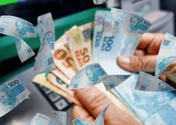 Dinheiro caindo de uma mão que segura notas de 100 e 50 reais, representando o recebimento do 13º salário.