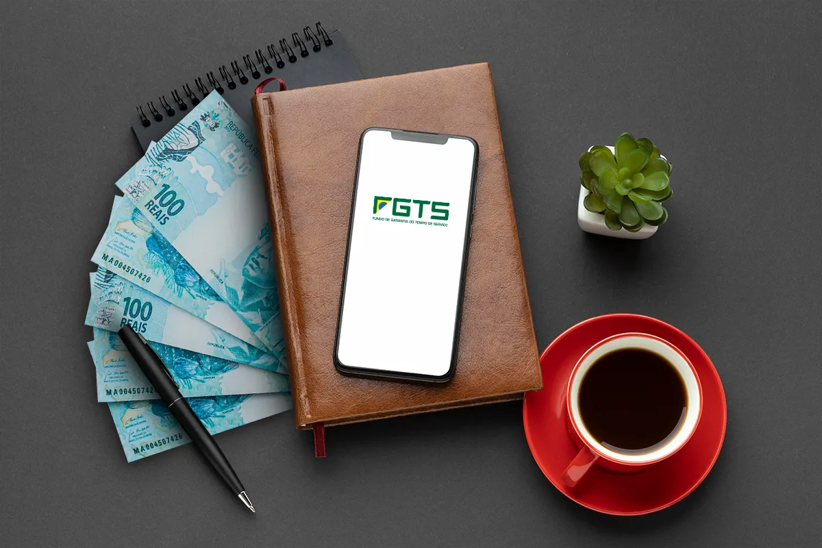 Smartphone com o logo do FGTS, cédulas de 100 reais, caderno, caneta e xícara de café, simbolizando o planejamento financeiro para a antecipação do Saque-Aniversário.