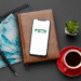 O app oficial do FGTS em um smartphone sobre uma mesa, junto a uma caneta, notas de reais e uma xícara de café. A imagem simboliza a retirada do saque-aniversário do FGTS.