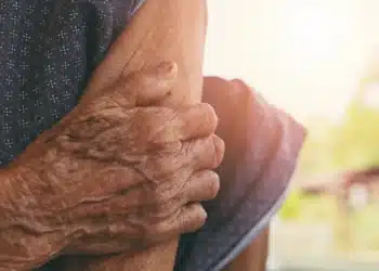 Mãos enrugadas de uma pessoa idosa segurando o braço, com luz solar ao fundo. Este texto é crucial para a acessibilidade, descrevendo a imagem para pessoas com deficiência visual e ajudando o Google a entender o conteúdo visual.