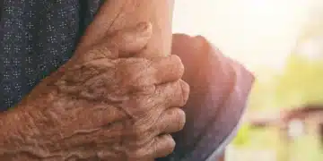 Mãos enrugadas de uma pessoa idosa segurando o braço, com luz solar ao fundo. Este texto é crucial para a acessibilidade, descrevendo a imagem para pessoas com deficiência visual e ajudando o Google a entender o conteúdo visual.