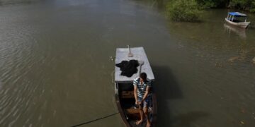 Pescador em sua embarcação em área fluvial, preparando redes de pesca, com vegetação ao fundo. Imagem ilustrando a prática da pesca artesanal.