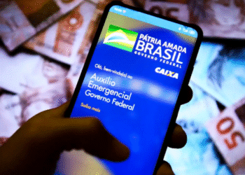 Tela de celular mostrando aplicativo do Auxílio Emergencial do Governo Federal, com fundo de cédulas de reais desfocadas.
