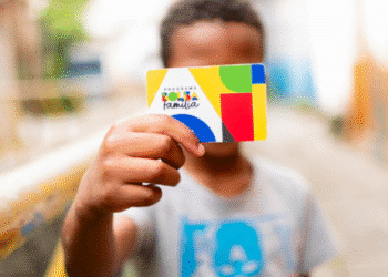 Criança negra sorridente segurando cartão colorido do Programa Bolsa Família em ambiente urbano ao ar livre.
