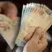 Mão segurando notas de real, representando antecipação do 13º salário para resolver dívidas antes do fim do ano.