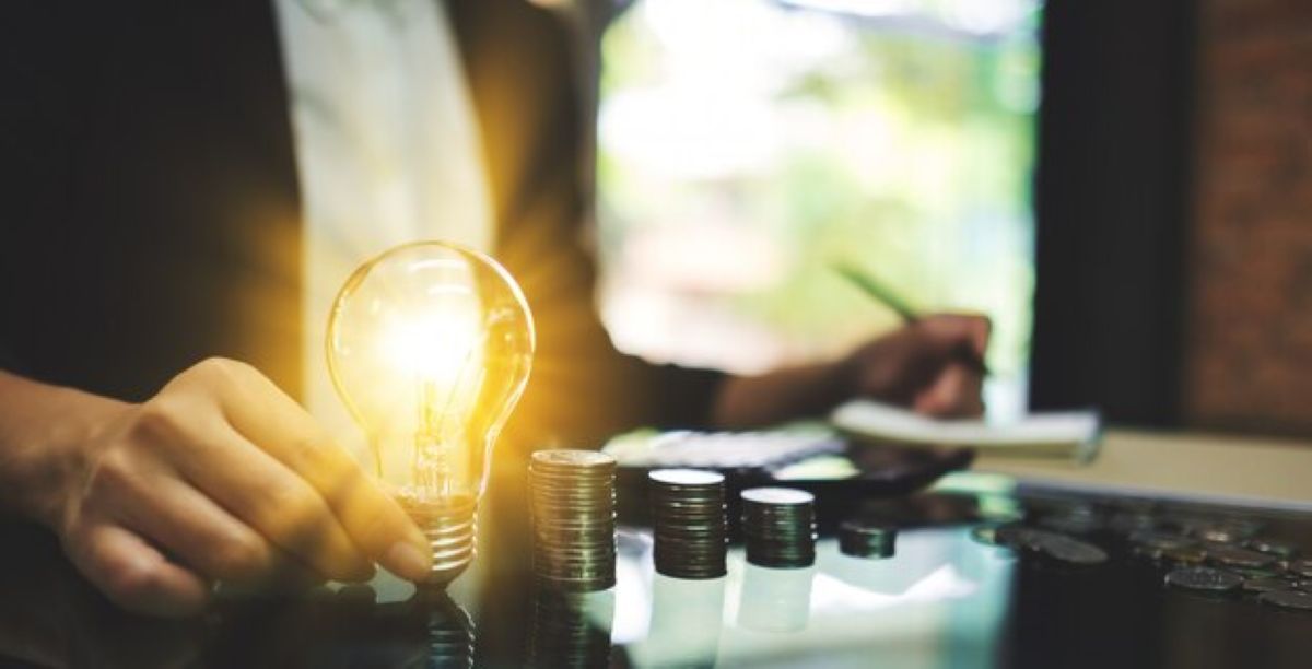 Pessoa segurando lâmpada acesa ao lado de moedas empilhadas representando economia de energia.