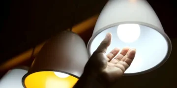 Mão tocando lâmpada de LED brilhante, com luz amarela e branca visíveis, ilustrando o conceito de energia elétrica em casa.