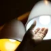 Mão tocando lâmpada de LED brilhante, com luz amarela e branca visíveis, ilustrando o conceito de energia elétrica em casa.