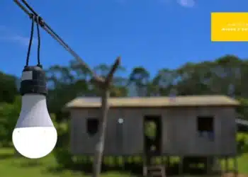 Imagem de uma lâmpada acesa em uma linha elétrica com uma casa ao fundo, simbolizando o programa Luz para Todos, que garante acesso à energia em áreas remotas.