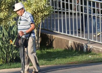 Um idoso usando chapéu e óculos escuros, caminhando com uma bolsa e um guarda-chuva em uma calçada, com grade de metal e vegetação ao fundo.