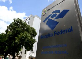 Placa da Receita Federal do Brasil sob céu azul com nuvens e árvores, simbolizando a instituição fiscal.