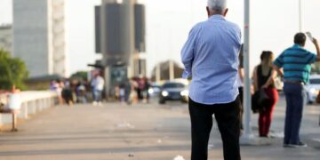 Homem idoso de costas em uma ponte movimentada, com pessoas e carros ao fundo, simbolizando a jornada da aposentadoria.