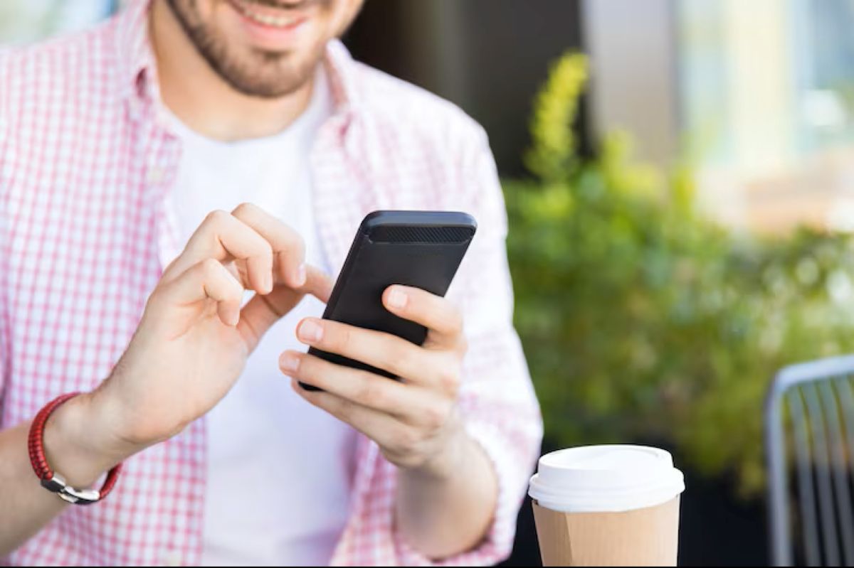 Homem sorrindo, usando o smartphone para navegar em um aplicativo, com um copo de café descartável na mesa.