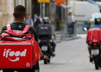 Entregador de aplicativo de delivery vestindo uma mochila grande e vermelha com o logo do iFood, pilotando uma moto em uma rua de cidade com tráfego.
