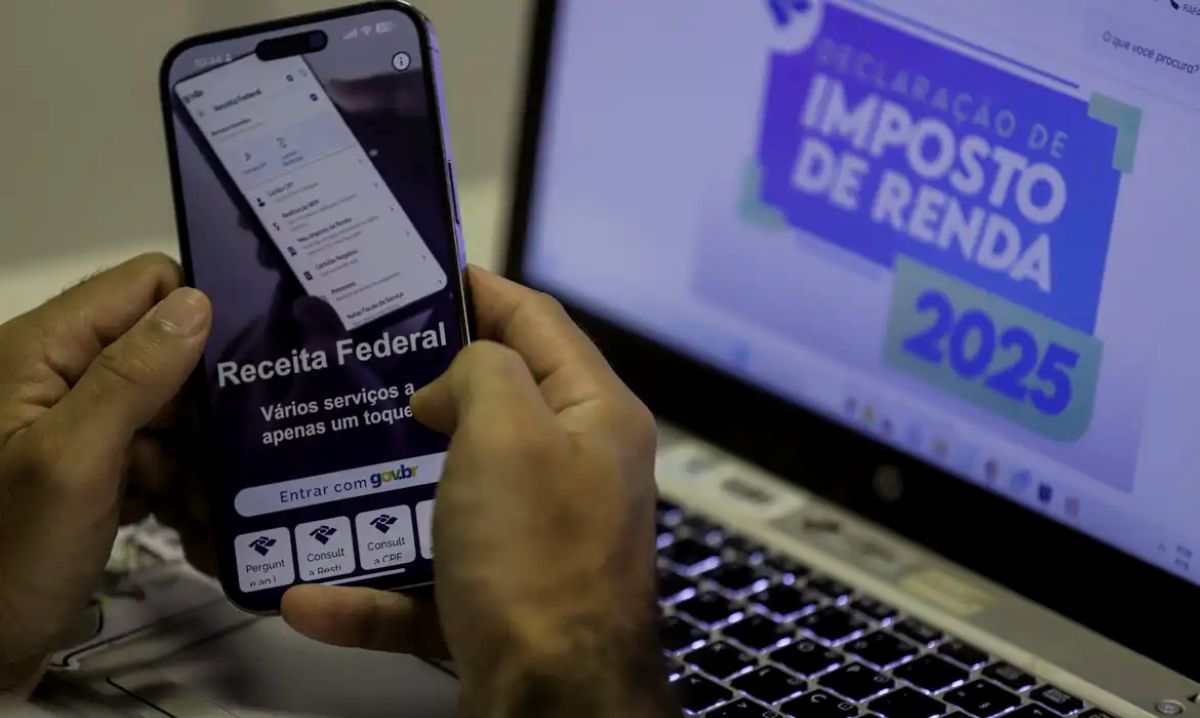 Pessoa usando o aplicativo da Receita Federal em um celular, com a tela de 'Declaração de Imposto de Renda 2025' em um notebook ao fundo.