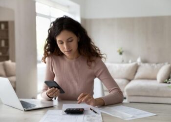 Uma mulher jovem sentada à mesa em casa, usando a calculadora do celular enquanto olha para alguns papéis e uma calculadora física.