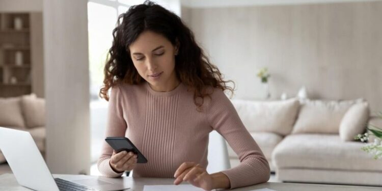 Uma mulher jovem sentada à mesa em casa, usando a calculadora do celular enquanto olha para alguns papéis e uma calculadora física.