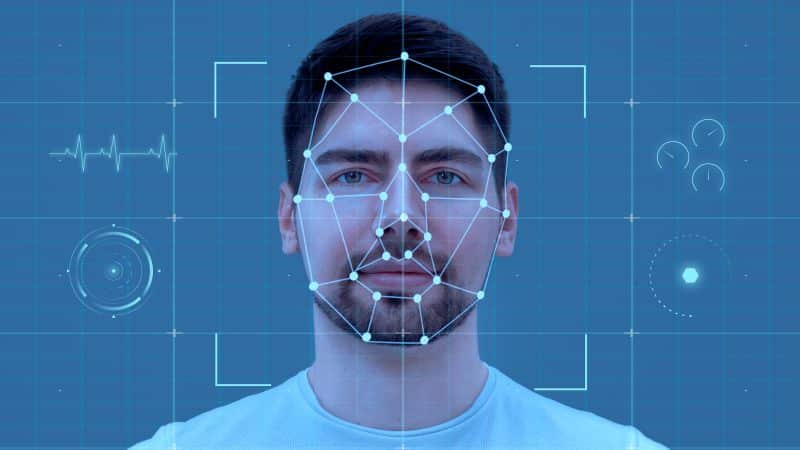 Um homem jovem com barba e uma mulher com cabelos encaracolados. O rosto de ambos tem uma sobreposição digital com pontos e linhas que mapeiam a biometria facial, sobre um fundo azul.