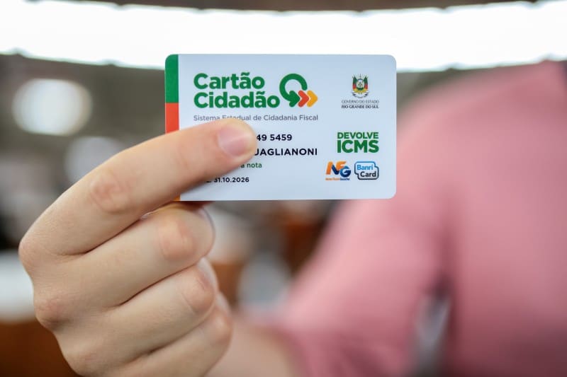 Mão segurando o Cartão Cidadão do programa Devolve ICMS, que representa o benefício trimestral. O cartão é a ferramenta para as famílias gaúchas acessarem o pagamento fixo de R$ 100,00, conforme o calendário de outubro de 2025.