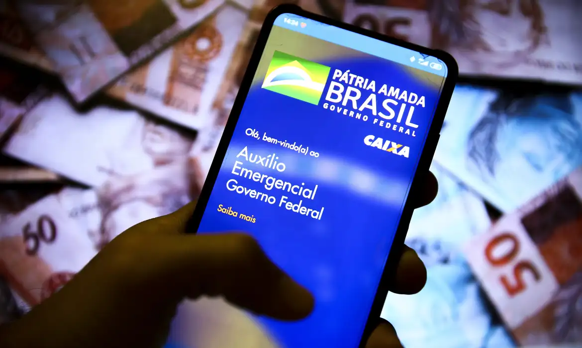 Smartphone exibindo o aplicativo "Auxílio Emergencial Governo Federal Caixa" sobre um fundo de notas de Real (R$ 50), representando a notificação do MDS para reaver R$ 478,8 milhões em pagamentos indevidos.