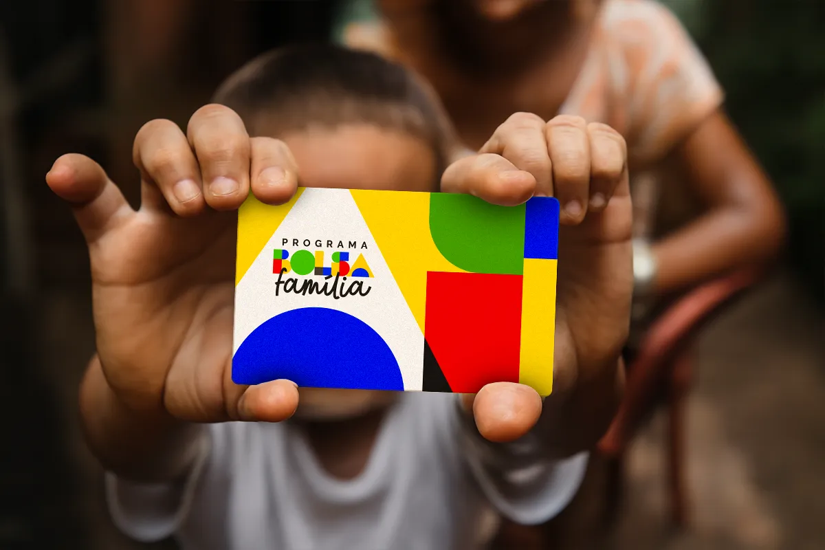 Cartão do Programa Bolsa Família com design colorido, em mãos de uma criança, representando a antecipação do pagamento do NIS final 6.